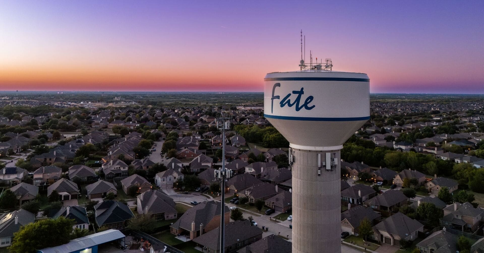 Fate, Texas water tower at sunset overlooking a suburban neighborhood with rows of residential homes and tree-lined streets.