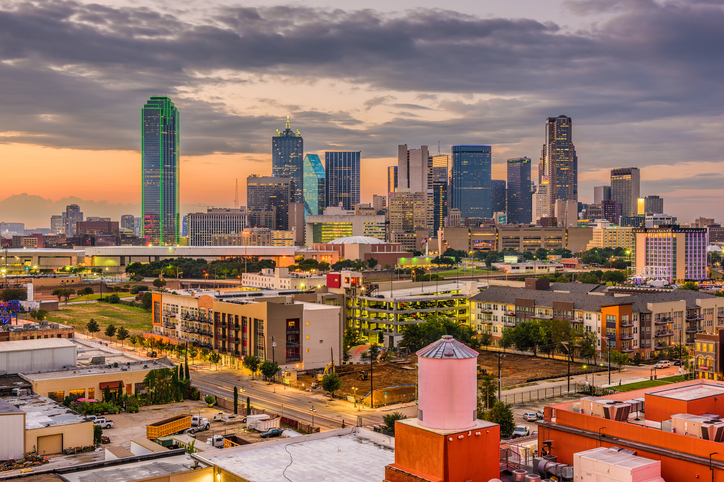 -A scenic sunset view of downtown Dallas, Texas, featuring the illuminated skyline with iconic skyscrapers like the Bank of America Plaza, modern high-rise buildings, residential complexes, and busy streets in the foreground. The image captures the urban energy and architectural diversity of the city.