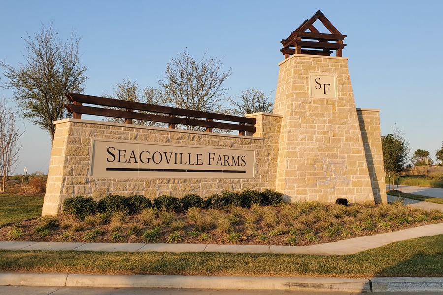 Stone monument sign at the entrance of Seagoville Farms community in Seagoville, Texas, featuring well-maintained landscaping, a tan brick structure with "SF" initials, and the community name engraved on a beige plaque.
