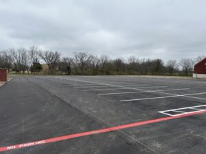 Freshly striped, empty asphalt parking lot with clear white parking lines and a bold red “No Parking” fire lane marking, bordered by trees under a cloudy sky.