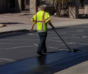 A construction worker in a high-visibility yellow shirt applies a fresh layer of sealcoat to an asphalt surface using a wide squeegee tool. The job is being done in a parking lot with visible parking space lines under bright daylight.