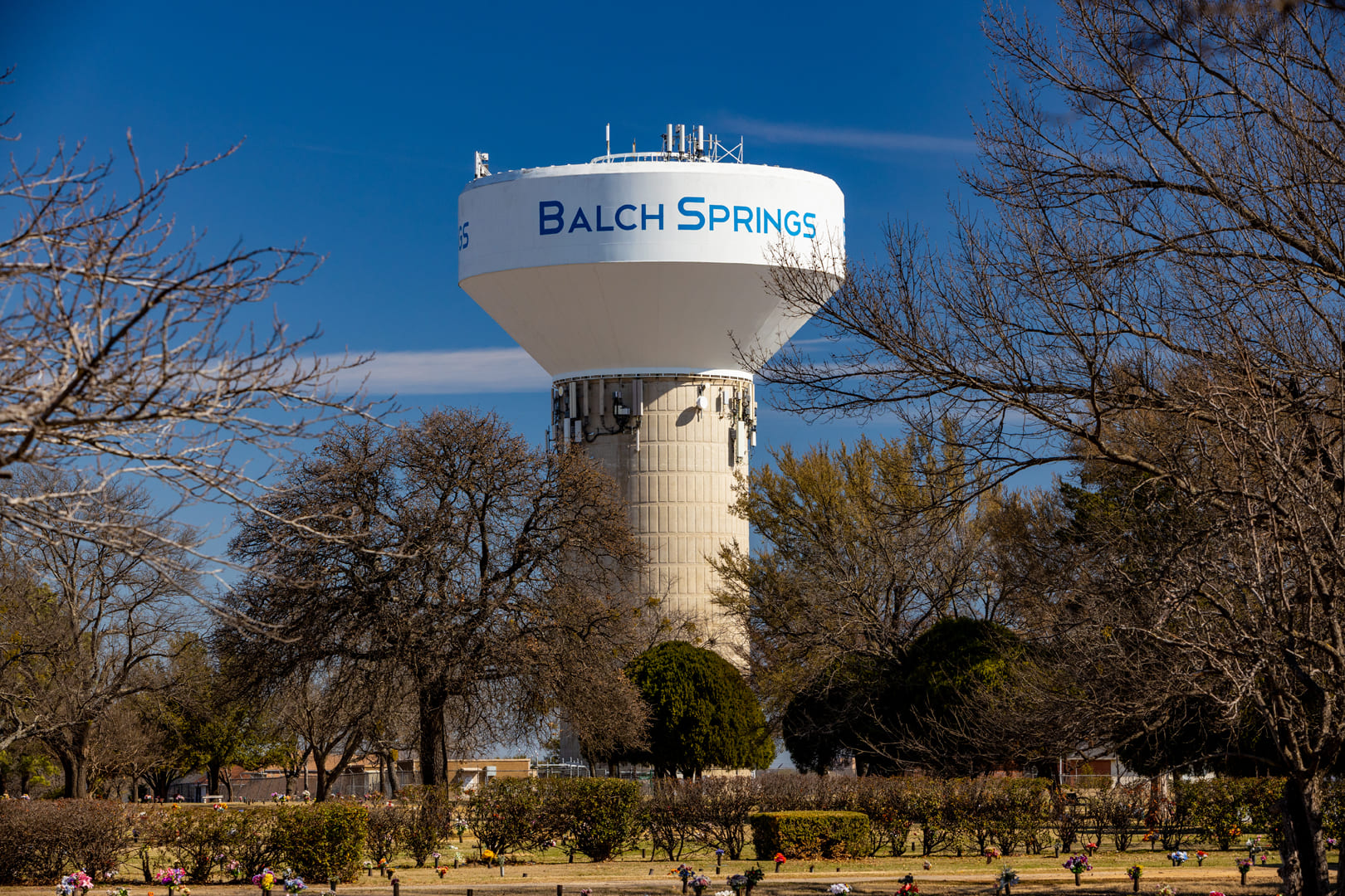 A tall water tower labeled "Balch Springs" stands prominently in a landscaped area surrounded by leafless trees and shrubbery under a clear blue sky, indicating a sunny winter day in Balch Springs, Texas.