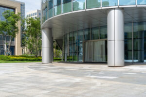 Modern commercial building entrance with large glass windows, silver structural columns, and a clean concrete plaza surrounded by greenery and landscaped office buildings in the background.