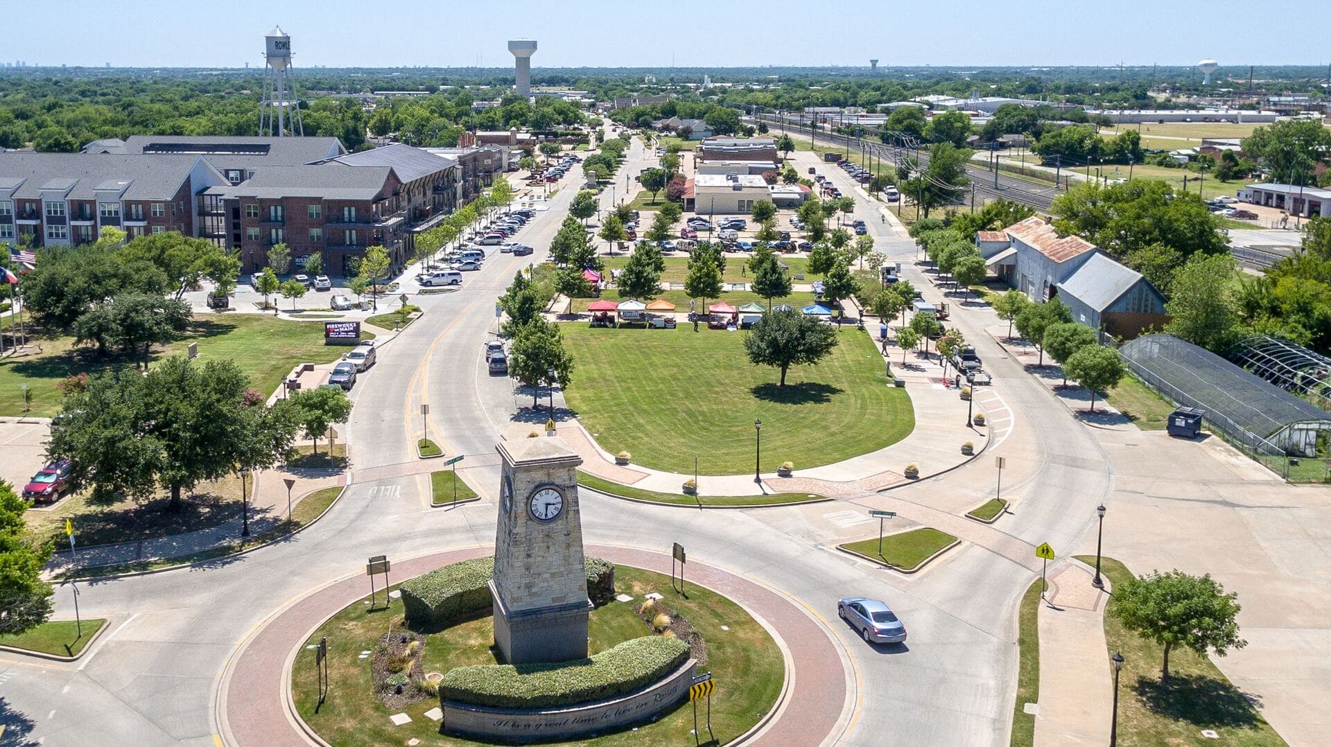Aerial view of Downtown Rowlett, TX showing a landscaped median with a central clock tower, flanked by shops, restaurants, and local vendors set up on a bright summer day, with surrounding roads and green trees.