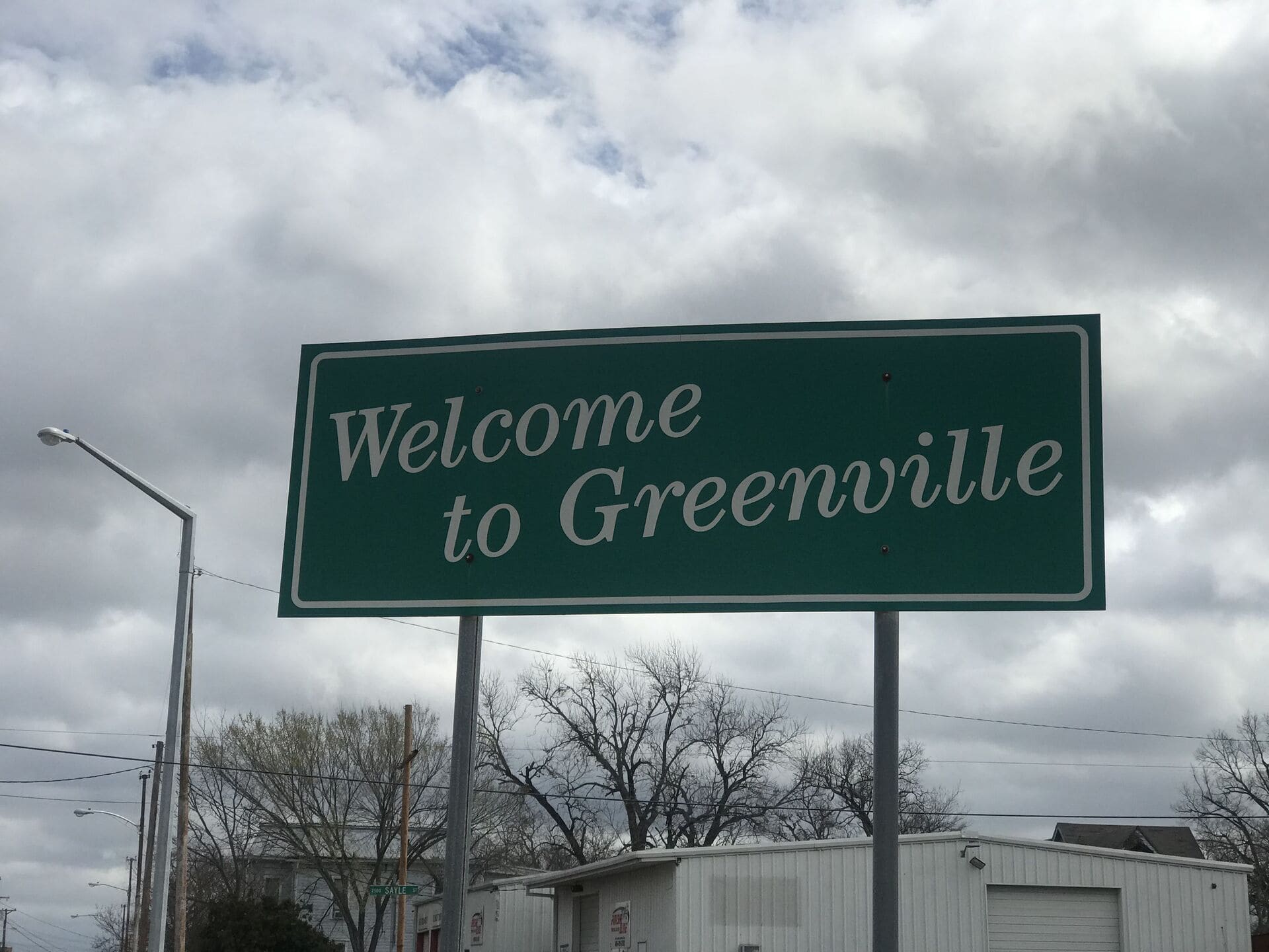Green road sign reading “Welcome to Greenville” with a cloudy sky and residential buildings in the background, taken in an urban area of Greenville, TX.