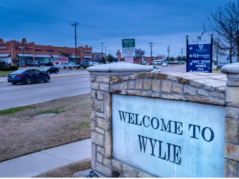 Stone and concrete 'Welcome to Wylie' sign at a roadside entry point in Wylie, Texas, with retail stores, traffic, and directional signage visible in the background under an overcast sky.