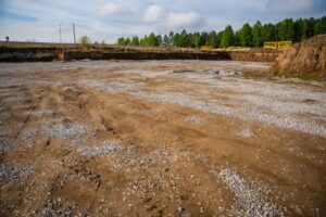 Construction site showing a prepared gravel base layer, ready for asphalt paving, with surrounding dirt embankments and forested area in the background.