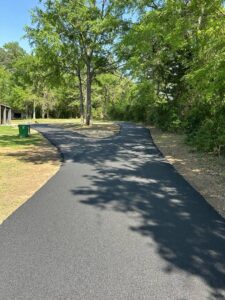 Freshly paved asphalt driveway curving through a wooded area in North Texas, surrounded by green trees and a clear blue sky.