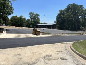 Asphalt roller compacting freshly paved roadway in a new commercial parking lot construction project in North Texas, surrounded by trees and curb installation.