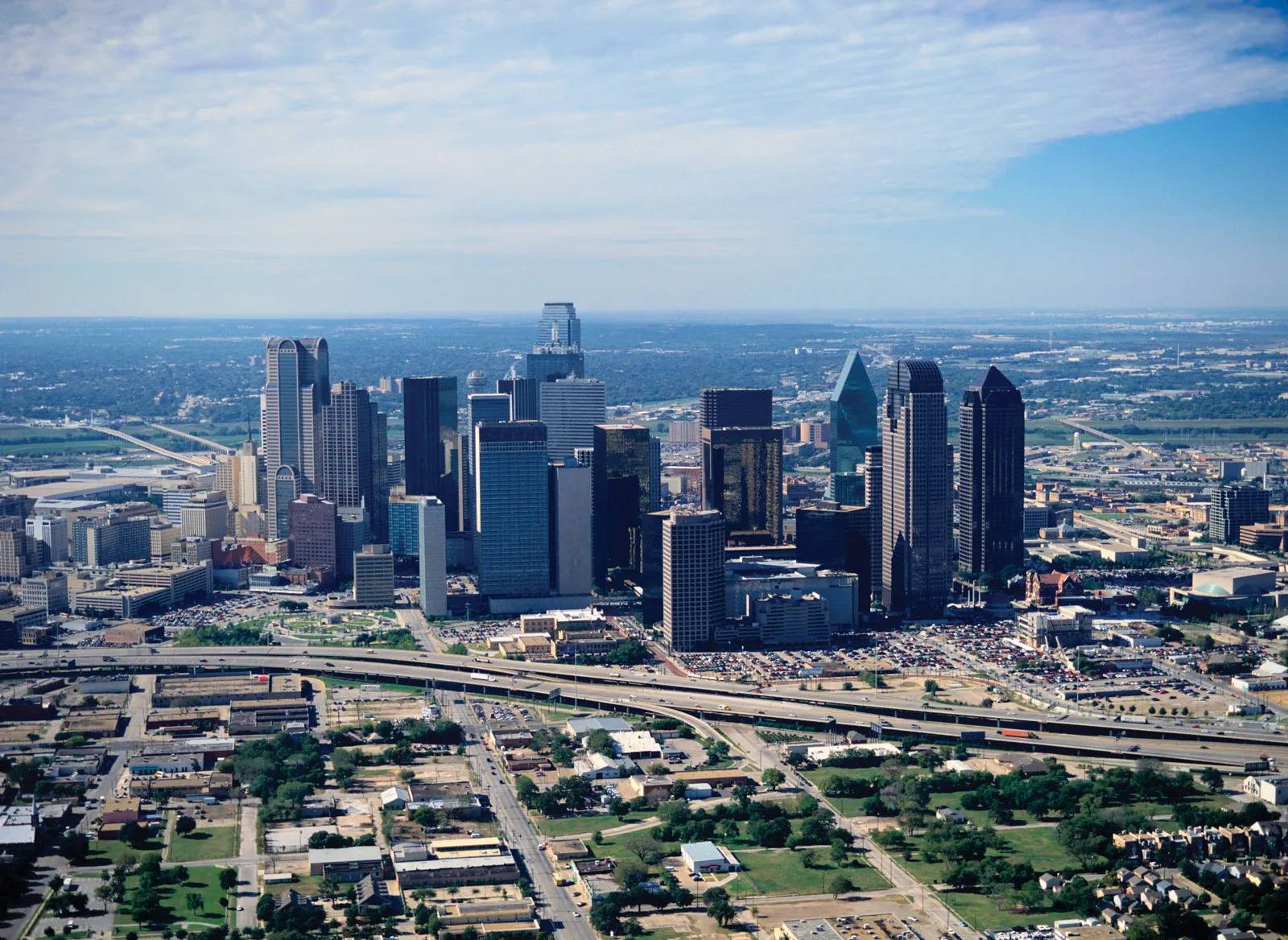 Aerial view of downtown Dallas, Texas, showcasing the skyline with high-rise buildings, highways, and surrounding neighborhoods under a partly cloudy sky.