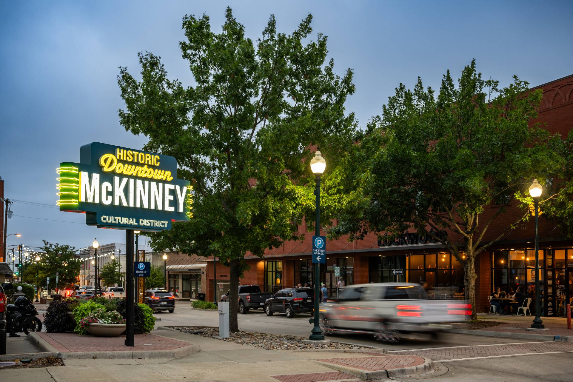 Historic Downtown McKinney, Texas street scene at dusk, featuring a lit sign, local shops, trees, and passing traffic.