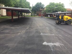 Freshly paved parking area being compacted with a roller near covered parking structures and residential buildings.