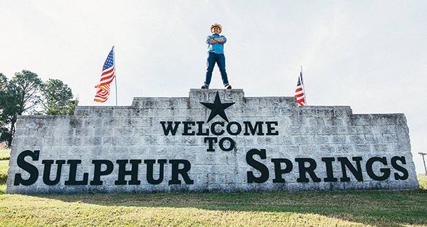 Welcome sign for Sulphur Springs featuring bold black lettering on a stone wall, with a statue standing on top and American flags on either side.