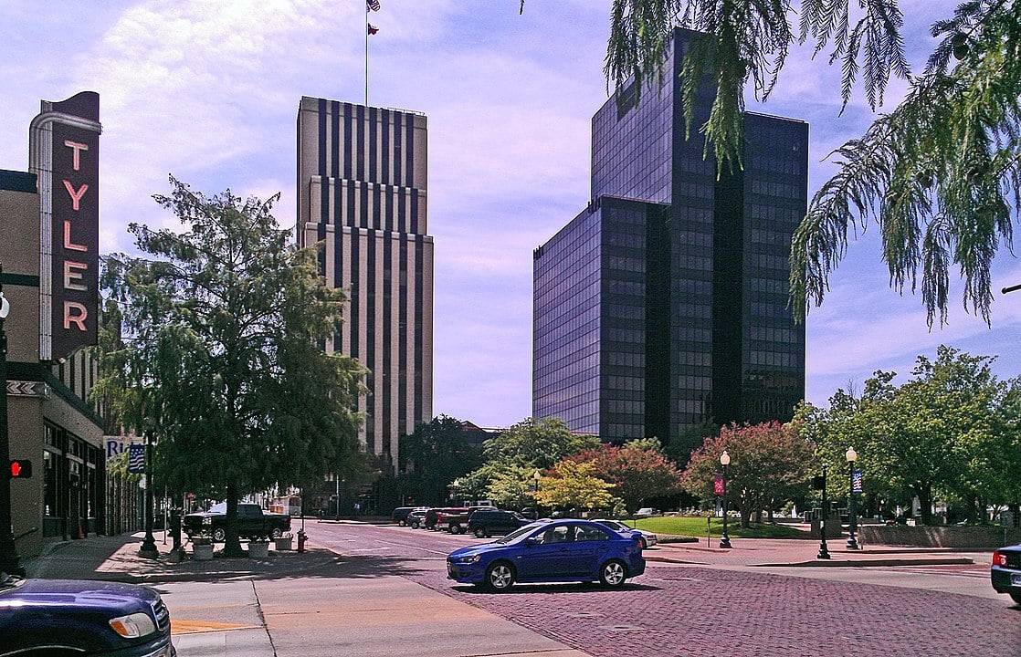 Downtown Tyler, Texas streetscape with mid-rise office buildings, a brick-paved intersection, trees, and a vertical “Tyler” theater sign along the sidewalk.