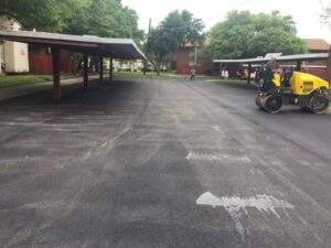 Freshly paved asphalt parking lot with a roller compactor in operation, covered parking structures on both sides, and residential buildings in the background.