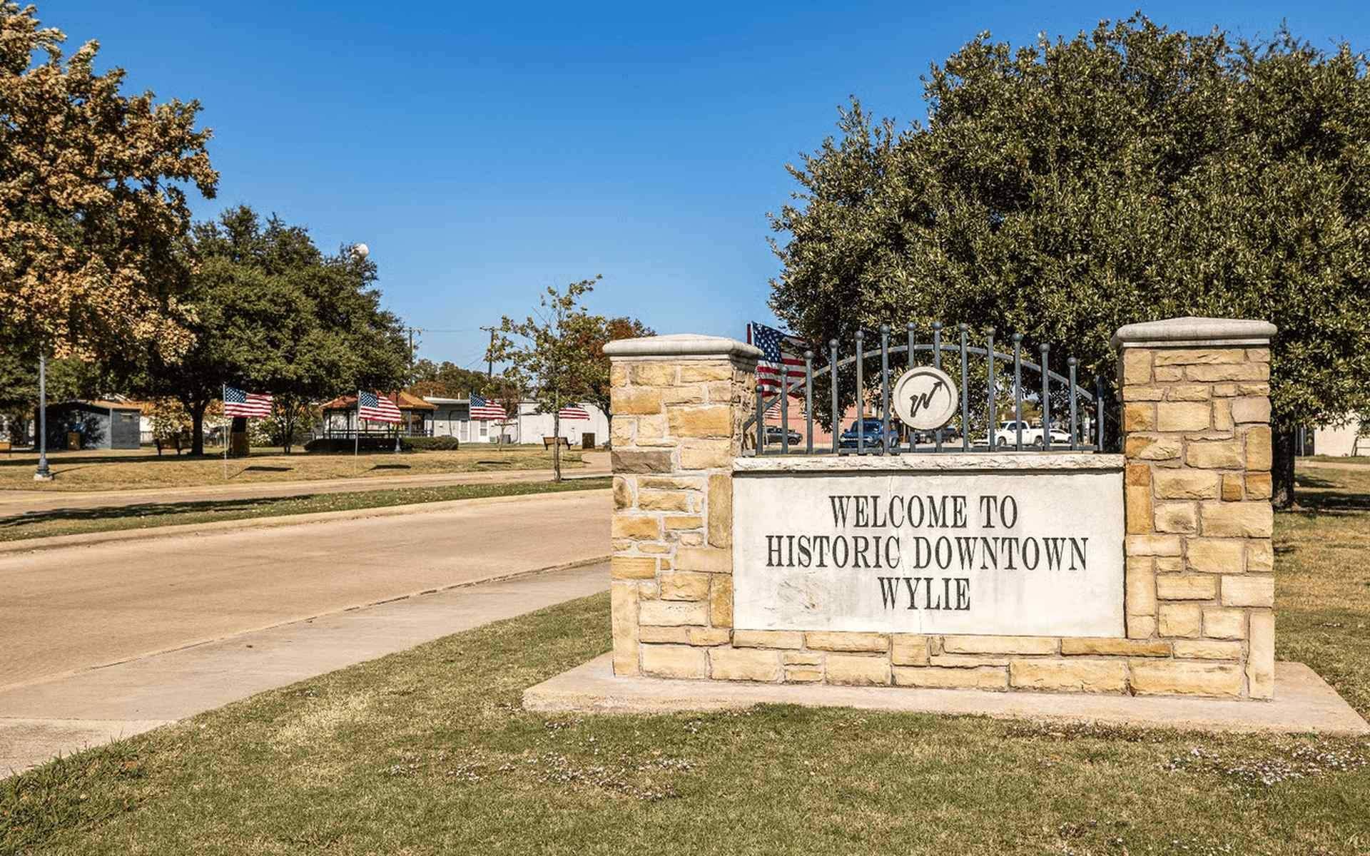 Stone entrance sign reading “Welcome to Historic Downtown Wylie” along a quiet roadway, with trees, grass, and American flags lining the background on a clear day.