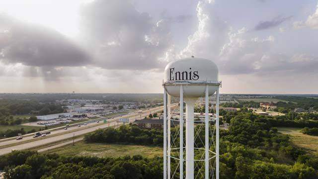 Aerial view of Ennis, Texas, featuring the city water tower overlooking a highway, commercial buildings, and surrounding green landscape beneath dramatic clouds.