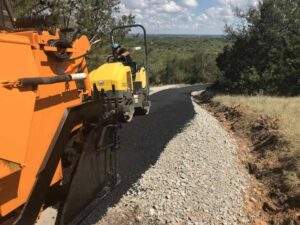 Asphalt paving crew upgrading a rural long residential driveway in summer, using heavy equipment to lay fresh pavement along a scenic, tree-lined landscape.
